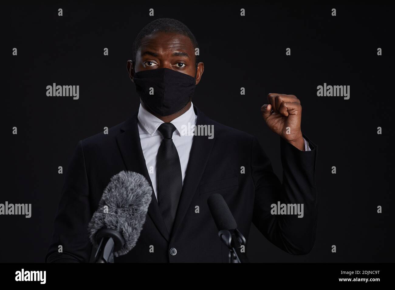 Portrait of African-American man wearing mask while giving powerful speech standing at podium against black background, copy space Stock Photo