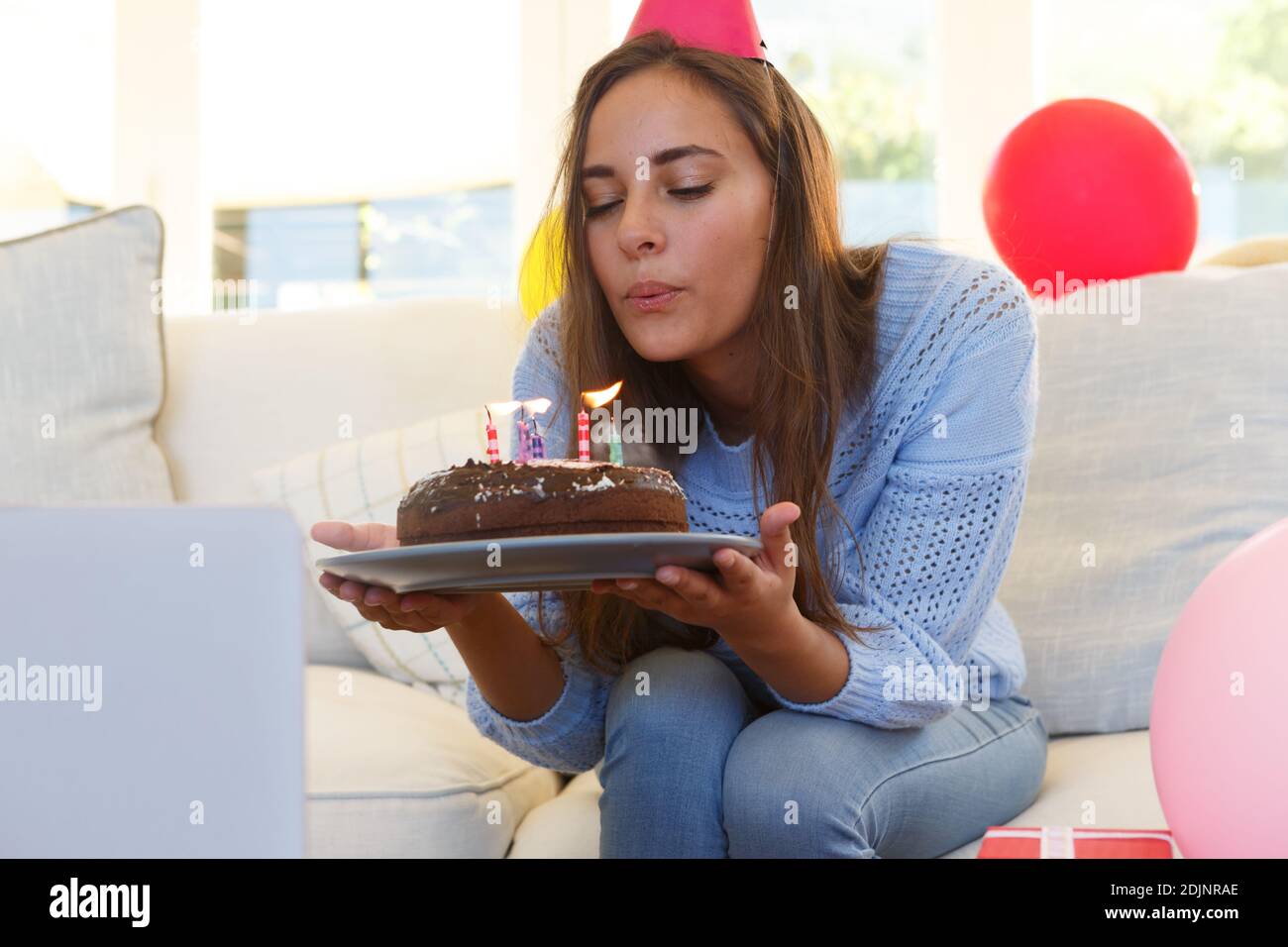 Caucasian woman having birthday video call blowing out candles on cake wearing party hat Stock Photo