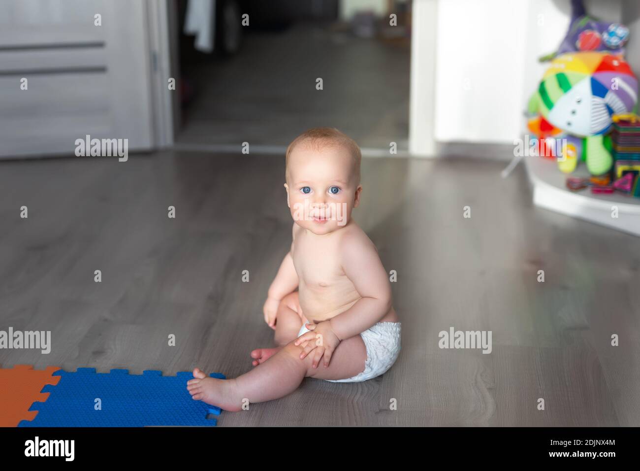 Cute adorable caucasian little infant baby boy enjoy playing in room sitting on floor with toys and soft mats on sunny day. Funny small child crawling Stock Photo