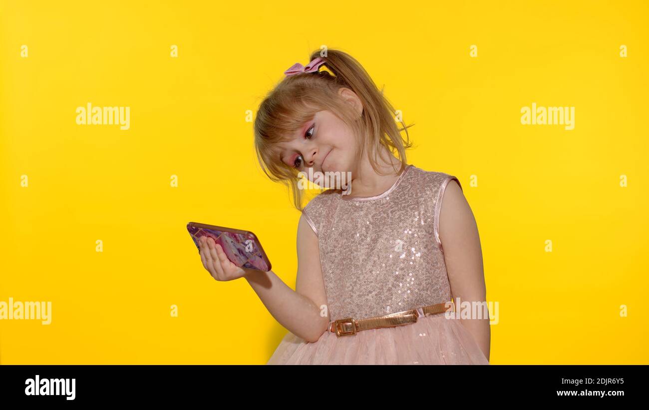 Child girl using smartphone. Portrait of kid with blonde hair look at the screen of a mobile phone, watch cartoons. Video call, blog, play games, looking at cellphone. Yellow background. Stock Photo