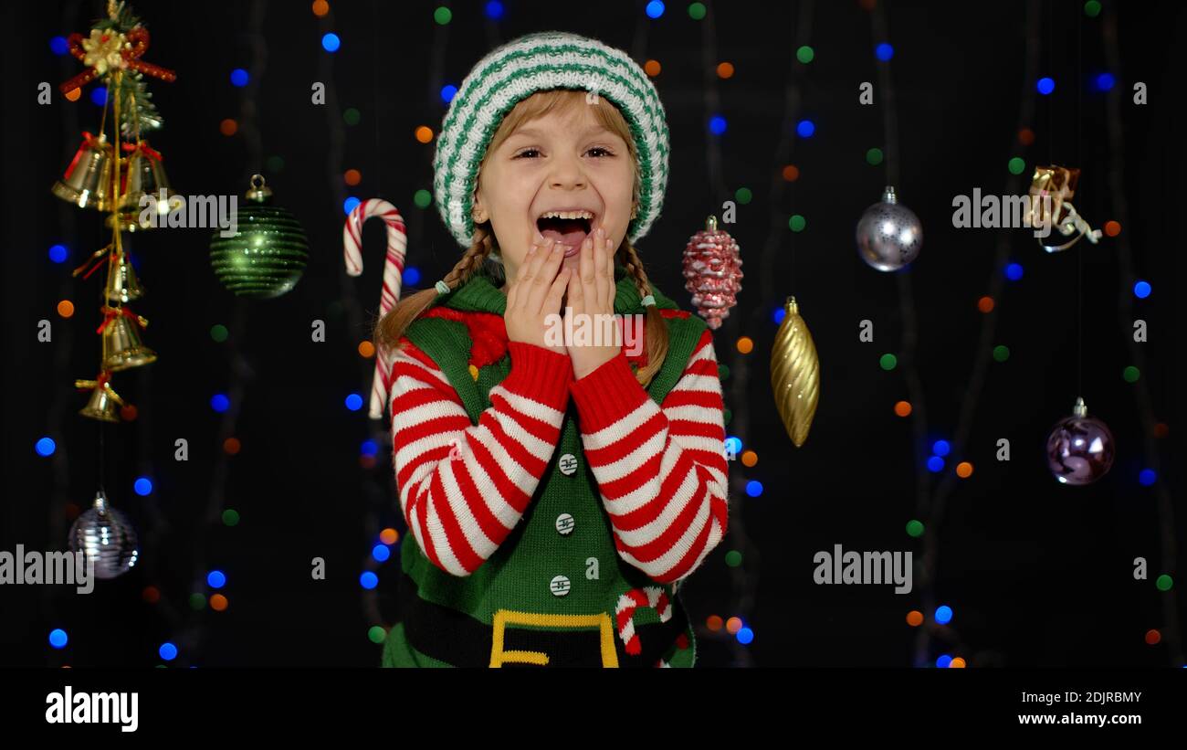 Overjoyed teen kid girl in Christmas elf Santa Claus helper costume laughs happily hears funny anecdote isolated on black background with garland. Child cannot stop laughing. Holidays celebration Stock Photo