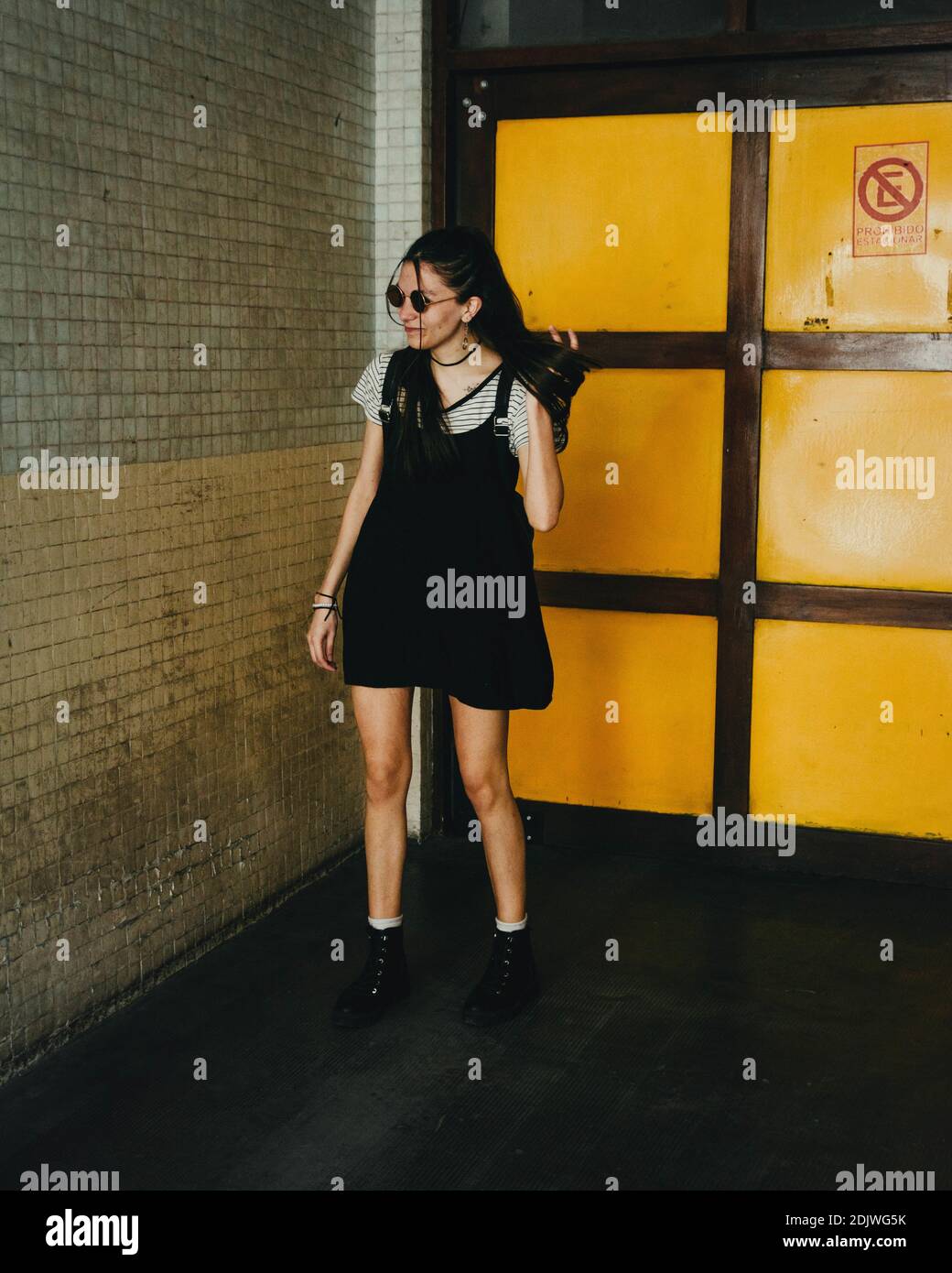 A vertical shot of a beautiful Caucasian girl with a black dress and sunglasses posing in the hallway Stock Photo