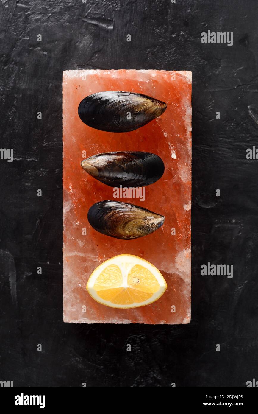 Seafood cooking on Himalayan pink salt. Mussels in shells with lemon served on whole salt block on black background top view copy space Stock Photo