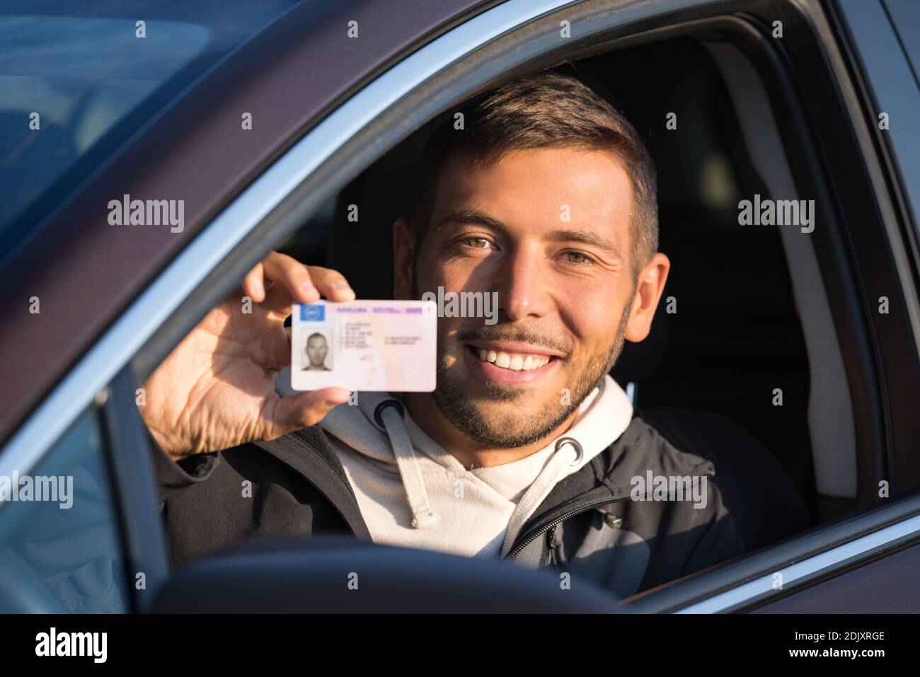 Attractive smiling young man proudly shows his driving license out of car window Stock Photo