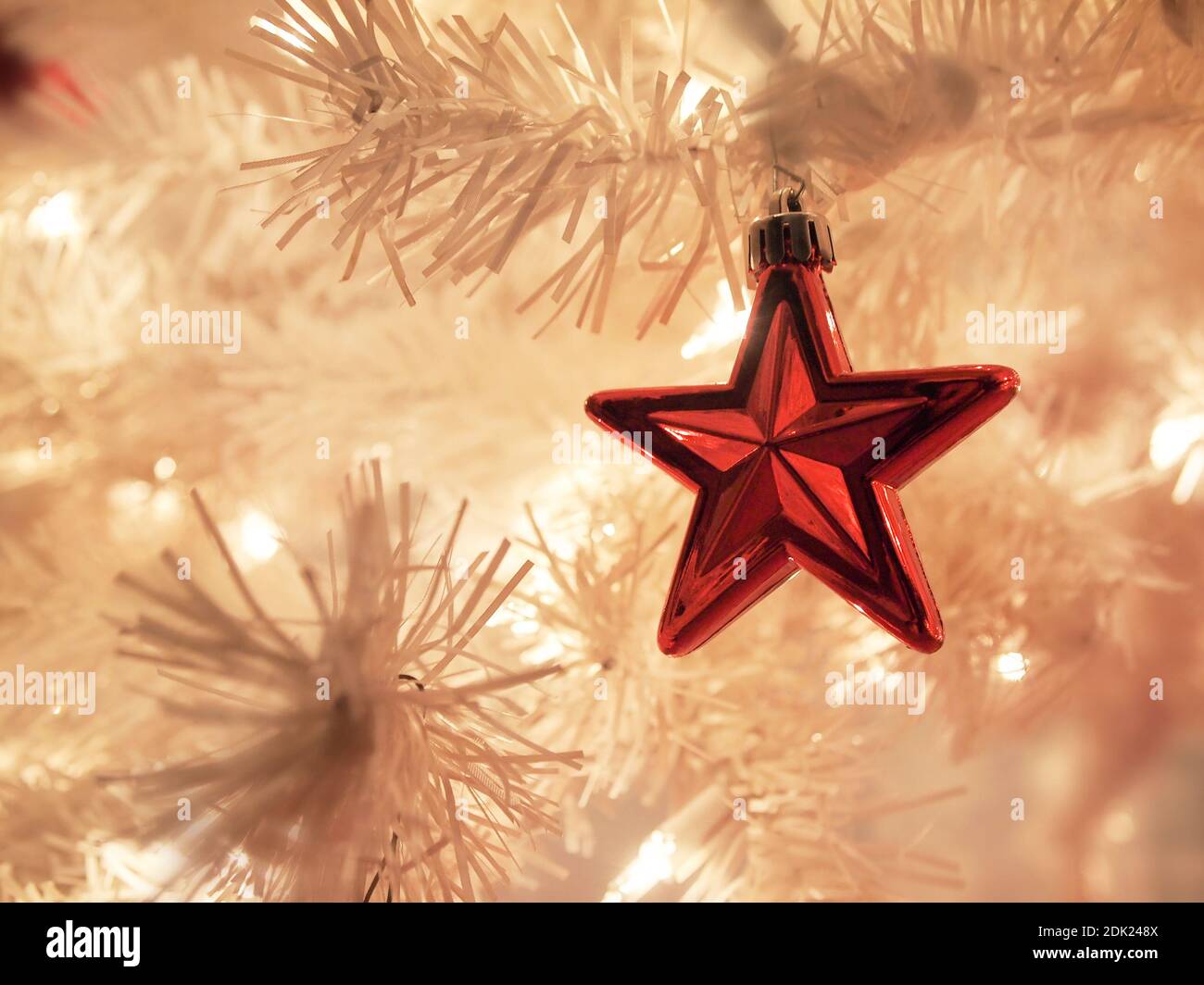 A shiny red star shaped ornament hangs in a white Christmas tree with bokeh and white twinkle lights during the holiday season. Stock Photo