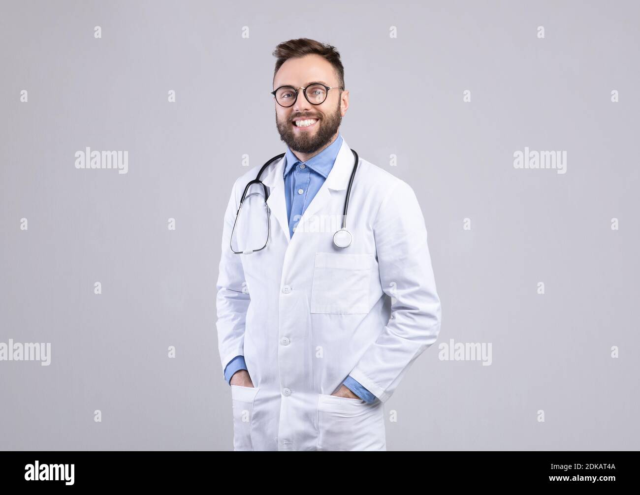 Confident Caucasian male doctor in white uniform smiling and posing over grey studio background Stock Photo