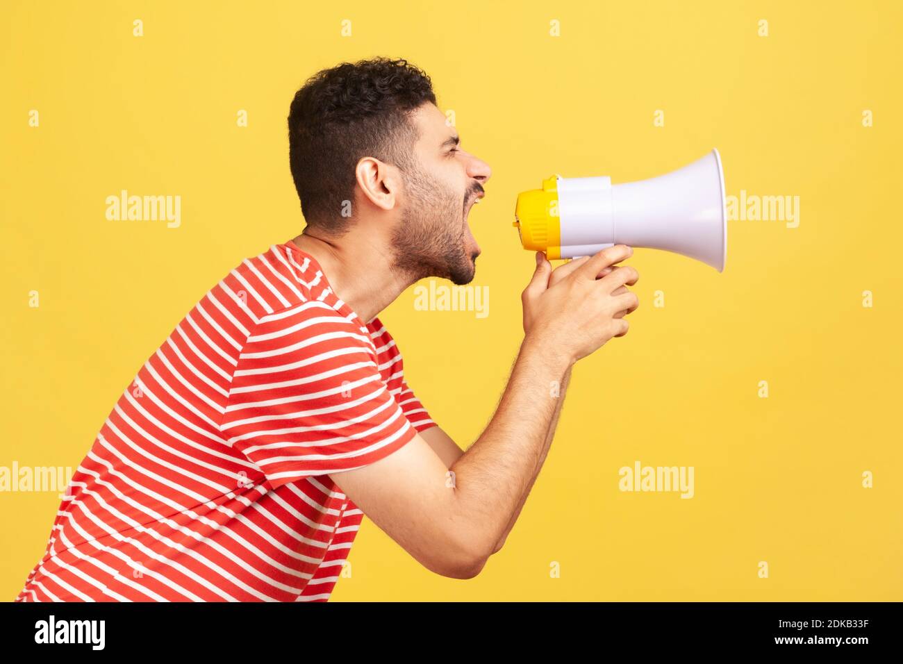 Profile portrait nervous bearded man in striped t-shirt loudly speaking screaming holding megaphone, announcing important message. Indoor studio shot Stock Photo