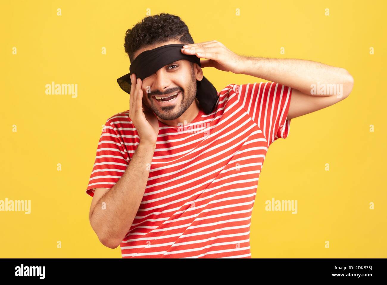 Happy bearded man in striped red t-shirt taking off blindfold from eyes to look at his surprise, looking at camera with toothy smile. Indoor studio sh Stock Photo