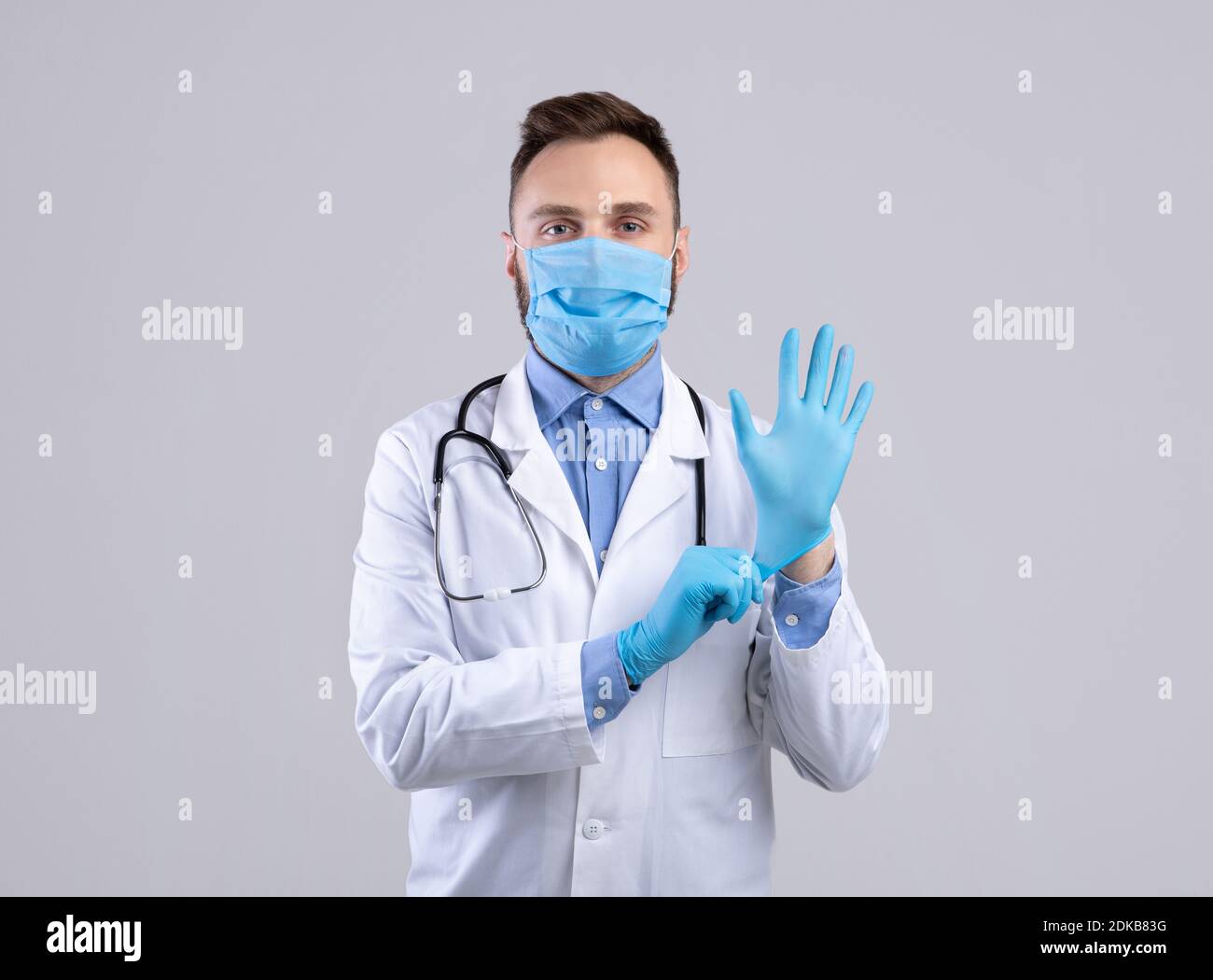 Confident young male doctor in facial mask putting on gloves over grey studio background Stock Photo