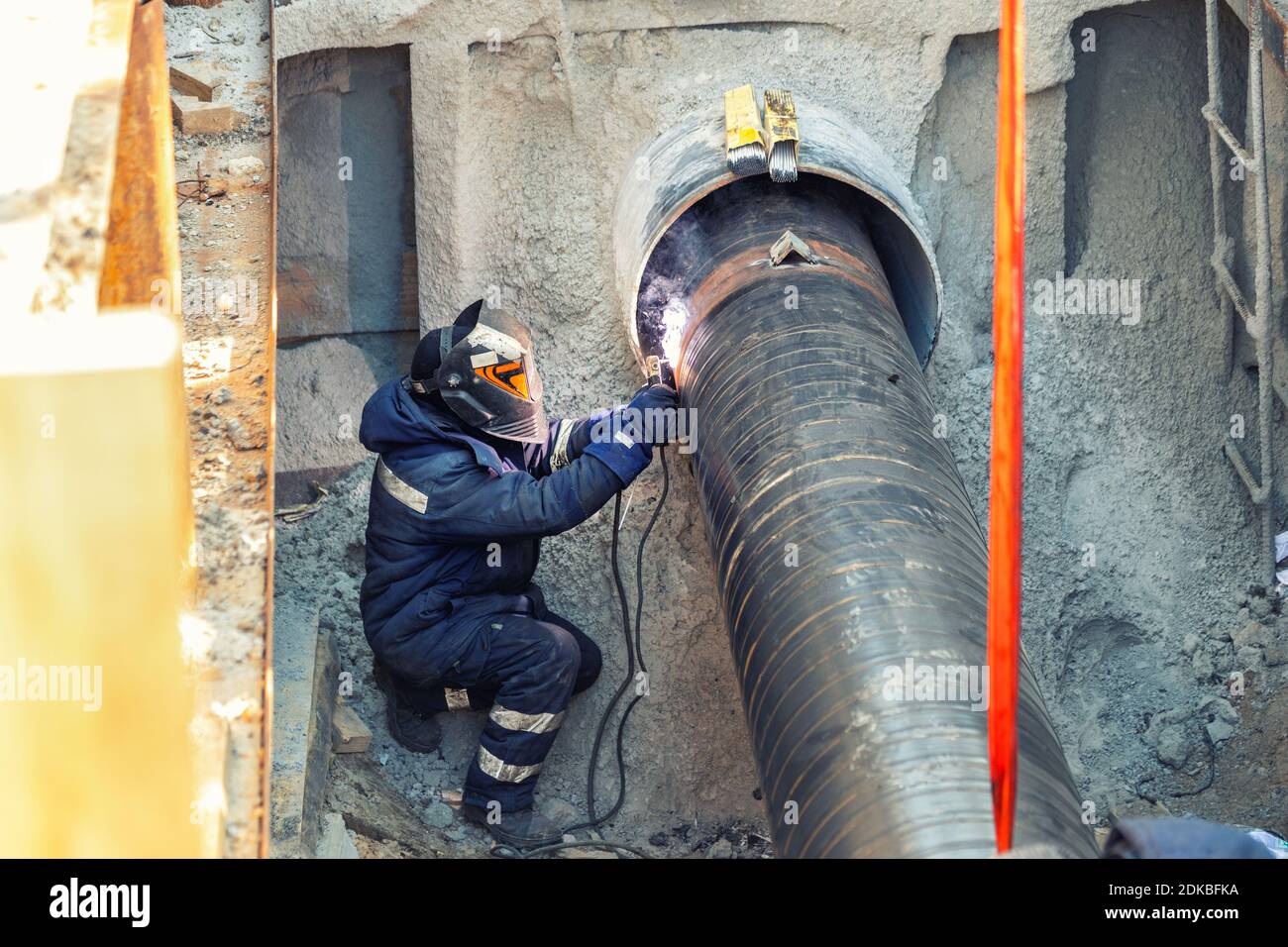 Professional welder welding water or gas steel pipeline in protective trench and mask at