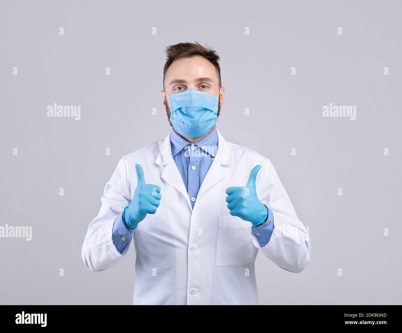 Millennial doctor in uniform, face mask and gloves looking at camera and showing thumbs up gesture over grey background Stock Photo