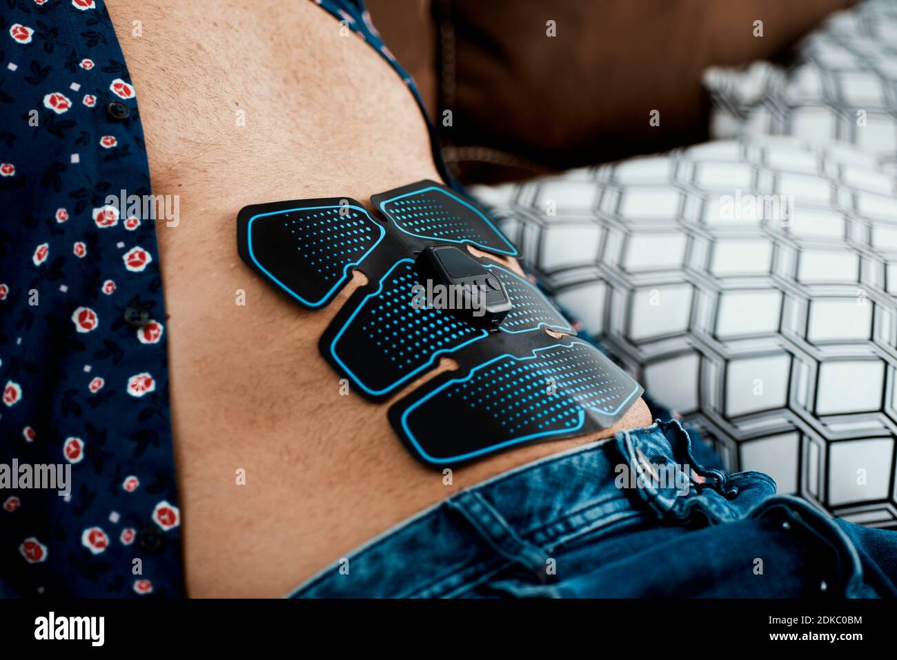 closeup of a young caucasian man using an electrical muscle stimulation device in his stomach while is sitting in a comfortable sofa Stock Photo