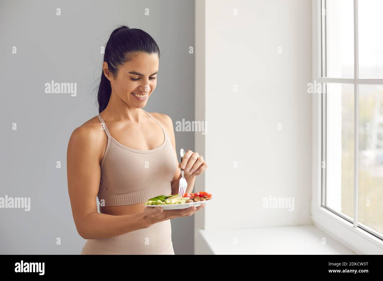 Happy fit woman enjoying healthy vegetarian meal, standing by the window at home Stock Photo