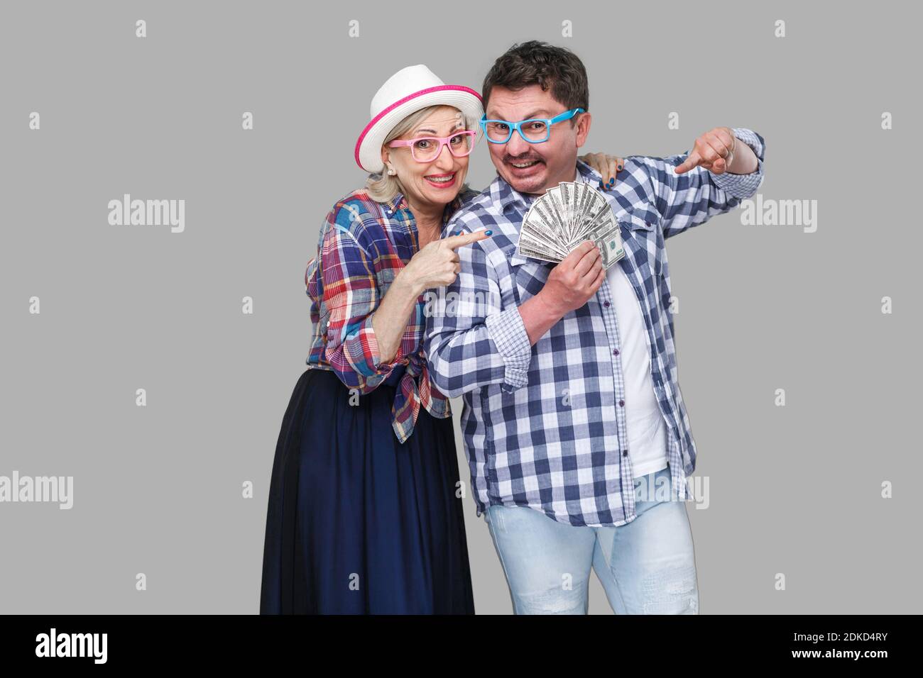Happy wealthy family, adult man and woman in casual checkered shirt standing pickaback together, holding fan of dollar and pointing finger to money. I Stock Photo