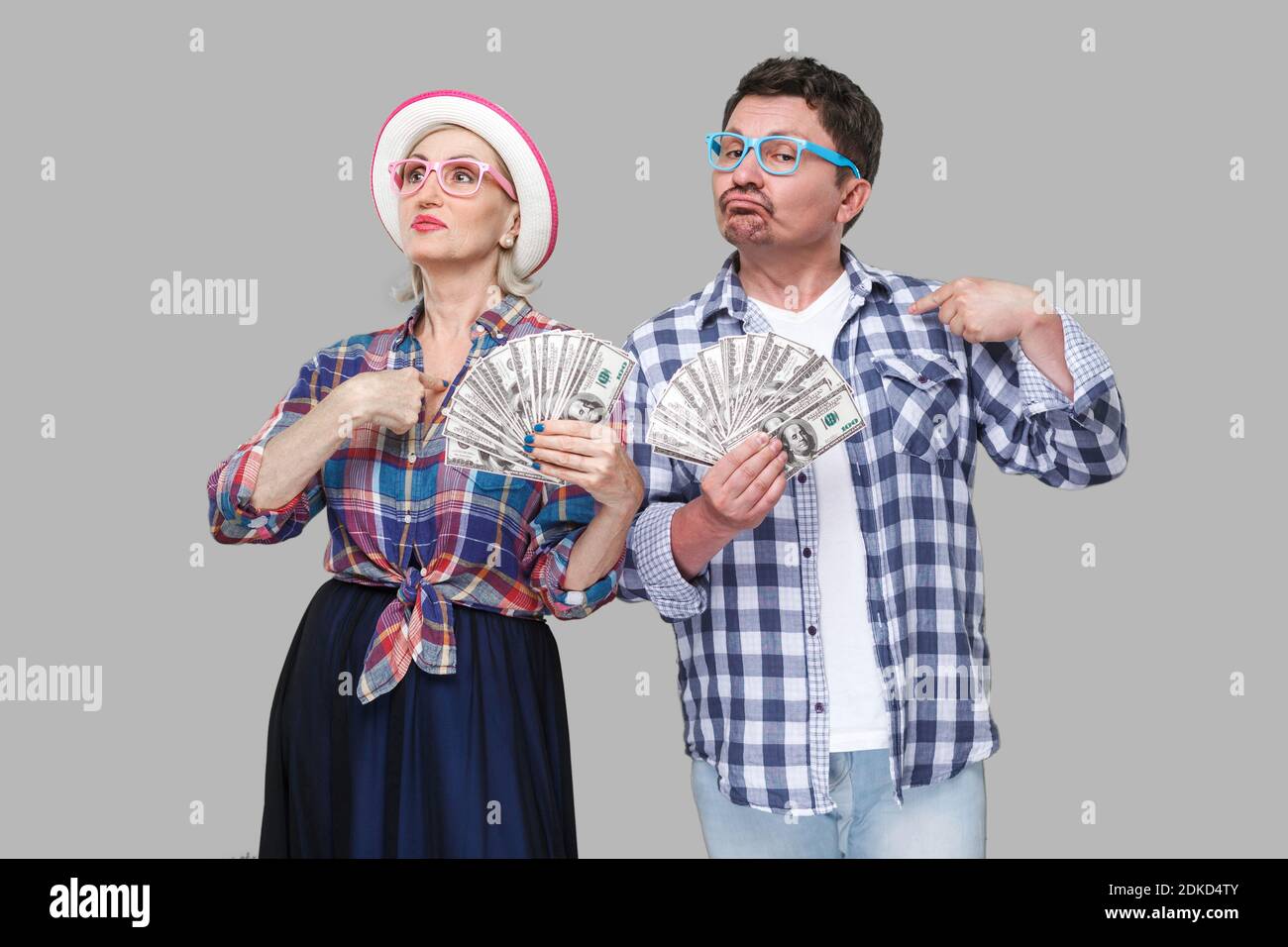 Couple of confident friends, adult man and woman in casual checkered shirt standing together holding fan of dollars and pointing fingers to themself w Stock Photo