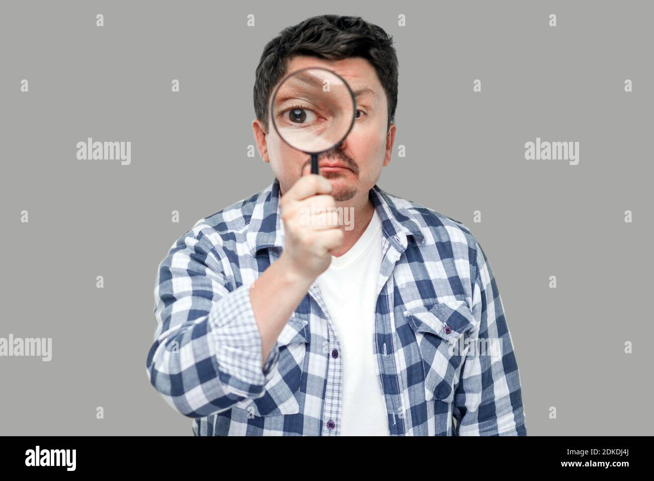 Portrait of serious middle aged man in casual checkered shirt standing, holding magnifying glass and looking at camera with big zoom eye. indoor studi Stock Photo