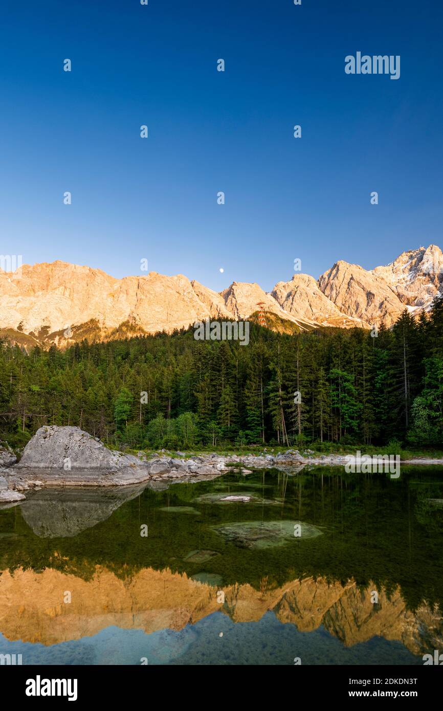 Reflection of the Wetterstein massif in the German Alps, during the evening light and three-quarter moon in a small lake by the Eibsee. In the background the Zugspitze. Stock Photo