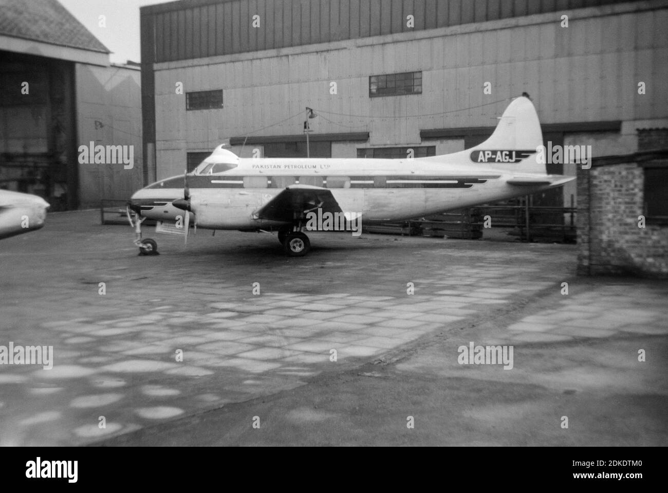 A vintage black and white photograph taken at London Gatwick Airport in 1967 showing a De Havilland Dove aircraft, registration AP-AGJ, belonging to Pakistan Petroleum Ltd. Aircraft is parked outside of a hangar. Stock Photo