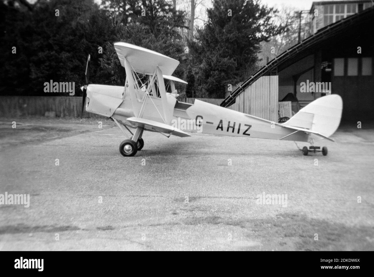 A vintage black and white photograph taken in 1954 showing a De Havilland Tiger Moth aeroplane, registration G-AHIZ, parked outside of a hangar in England. Stock Photo
