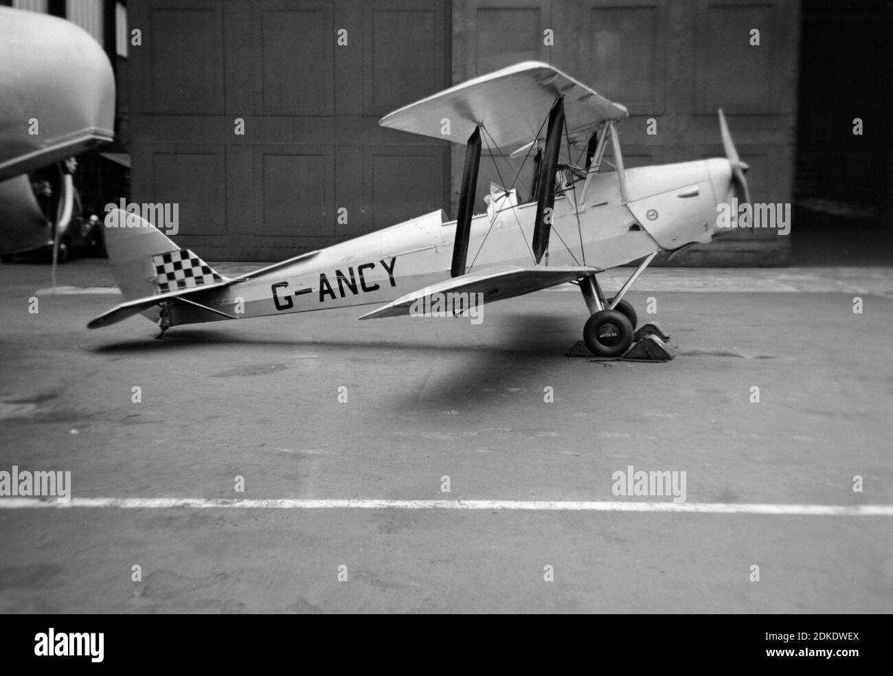 A vintage 1954 black and white photograph showing a De Havilland Tiger Moth aeroplane, registration G-ANCY, parked outside of a hangar. Stock Photo