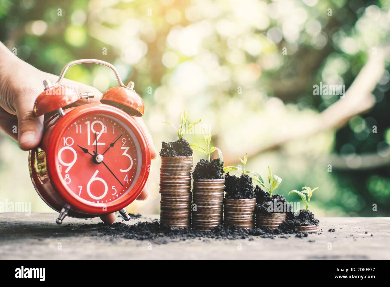 Cropped Image Of Hand Holding Alarm Clock By Stacked Coins And Piggy Bank On Table Stock Photo
