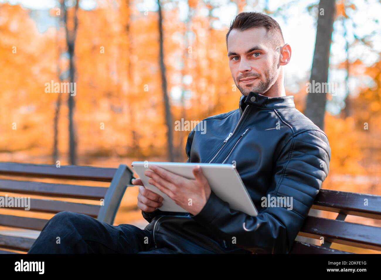 Handsome Young Man Sitting on the Bench and Using Big White Tablet PC at the Beautiful Autumn Park Stock Photo