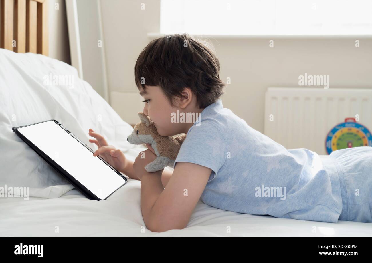 6-7 year old boy playing with dog toy and watching cartoon on tablet,Happy  child lying in bed playing game in the morning, Cute Kid having fun and  relaxing on his own in