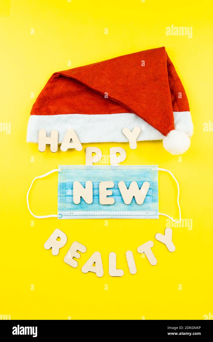 Santa hat, protective face mask and wooden letters HAPPY NEW REALITY on yellow background. Christmas and new year during epidemic concept. Stock Photo