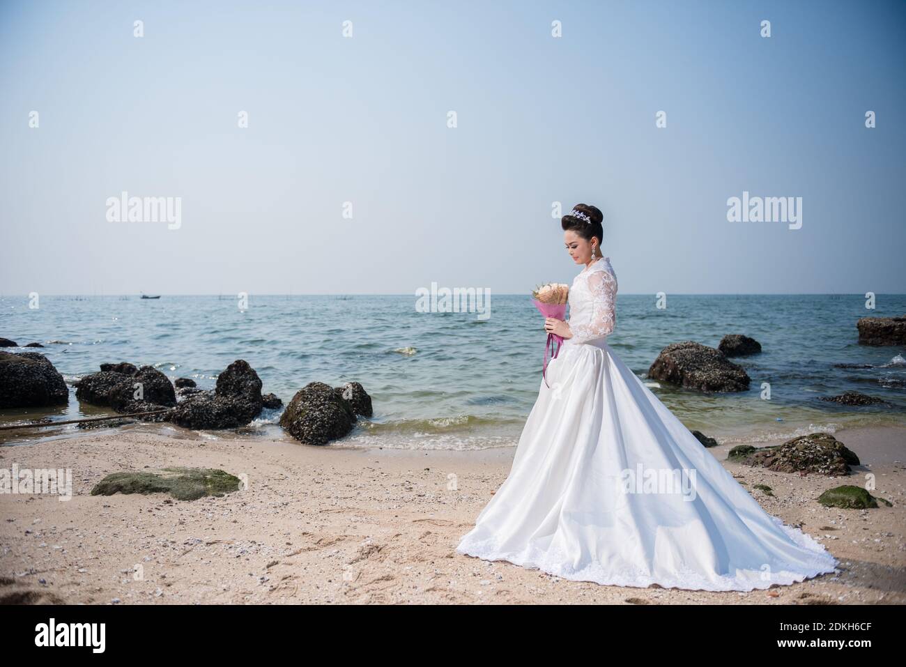 Beautiful woman in wedding dress holding flower standing on a beach ...
