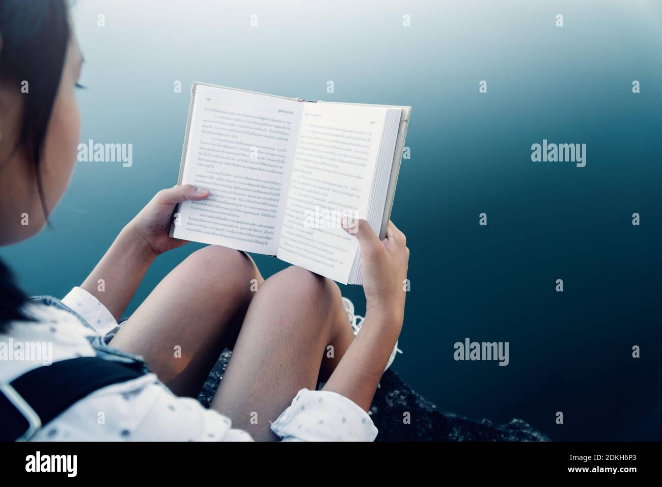 Midsection Of Girl Reading Book While Sitting Over Lake Stock Photo