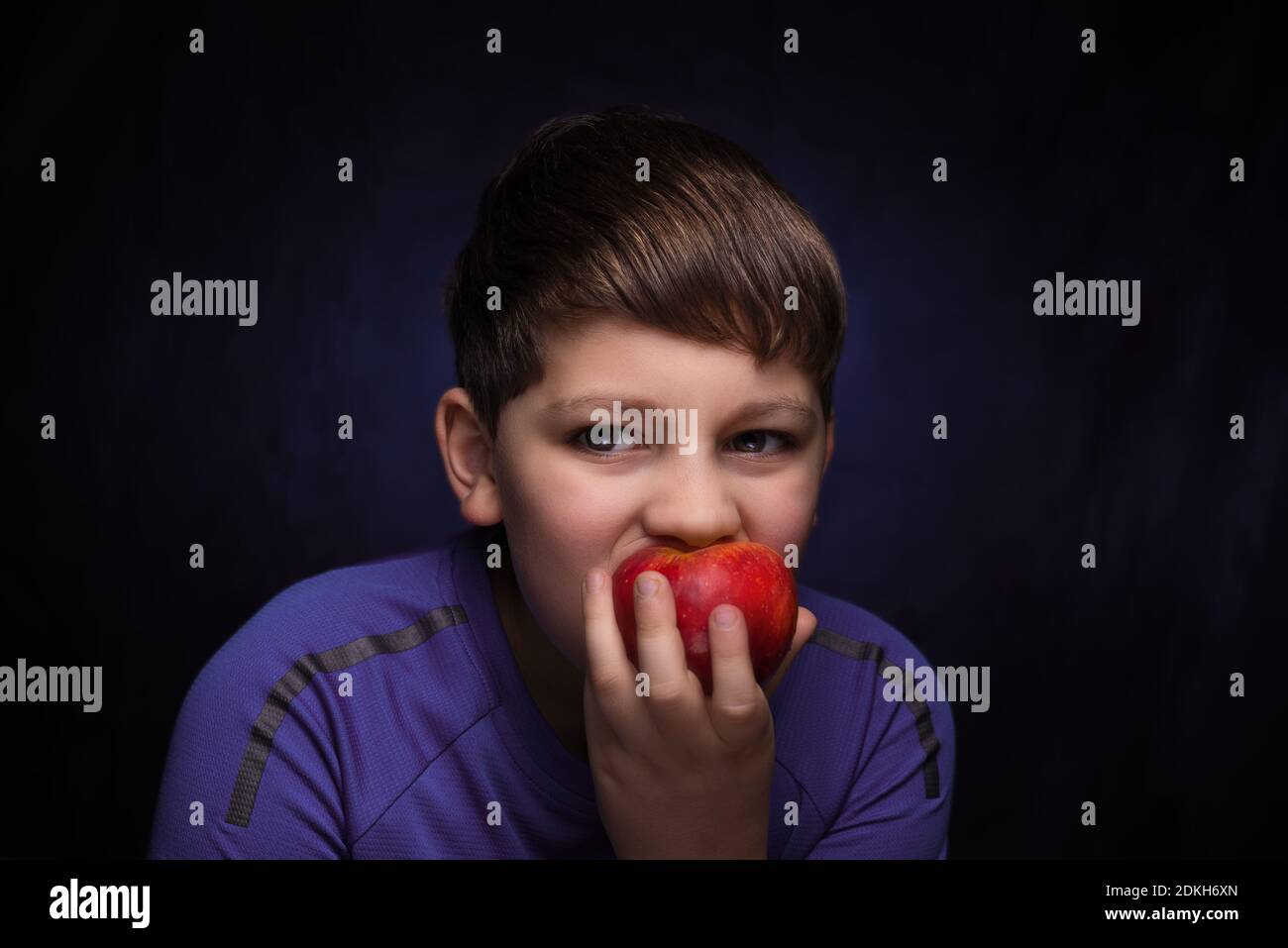 teenager boy with light brown hair and bangs, wearing a sports purple t-shirt eating a big red apple on an isolated background Stock Photo