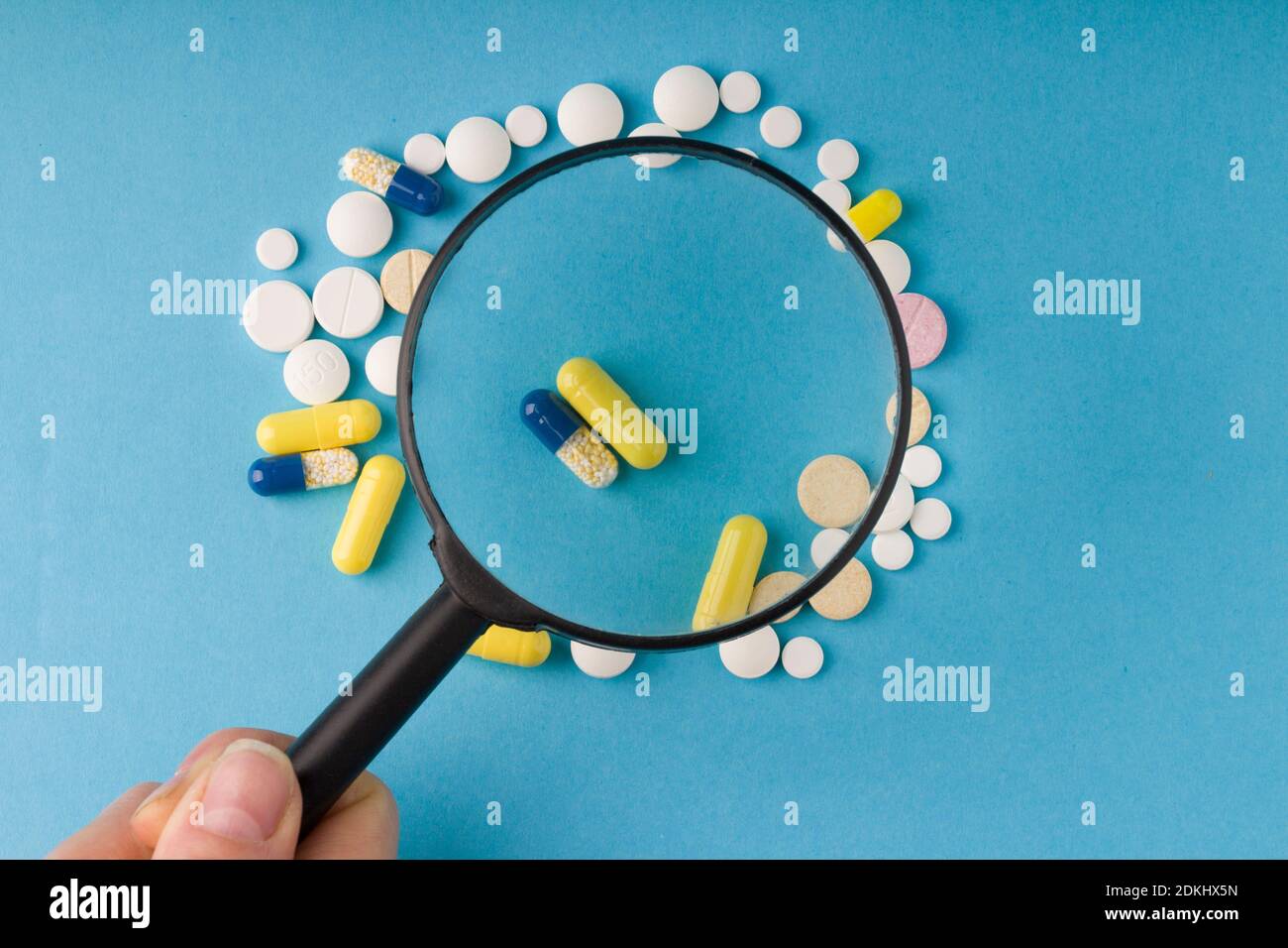 female hand holds a magnifying glass over pills, blue background . Concept of fakes of medicines and medicinal substance quality Stock Photo