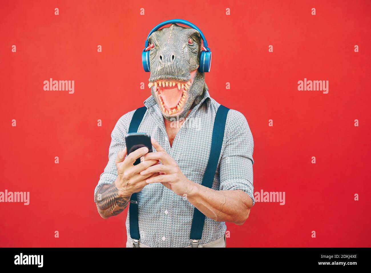 Senior fashion man wearing t-rex mask using mobile smartphone listening music with headphones - Crazy bizarre hipster guy having fun with new technology - Funny and absurd surreal concepts Stock Photo