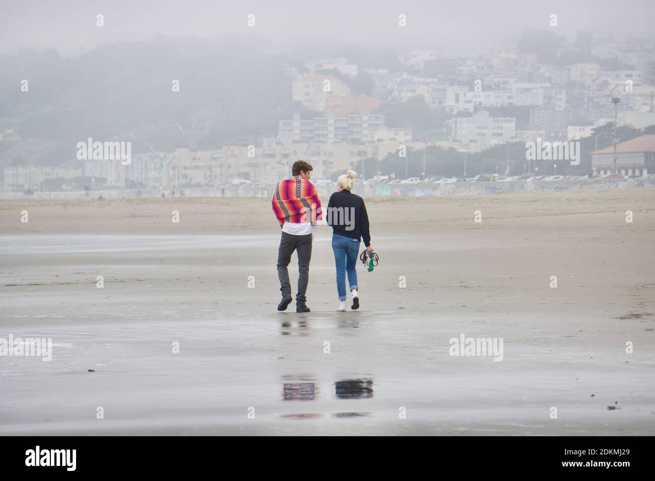 A couple walking on a cold, foggy Ocean Beach. Homes in the Richmond District in the background. San Francisco, California, CA Stock Photo
