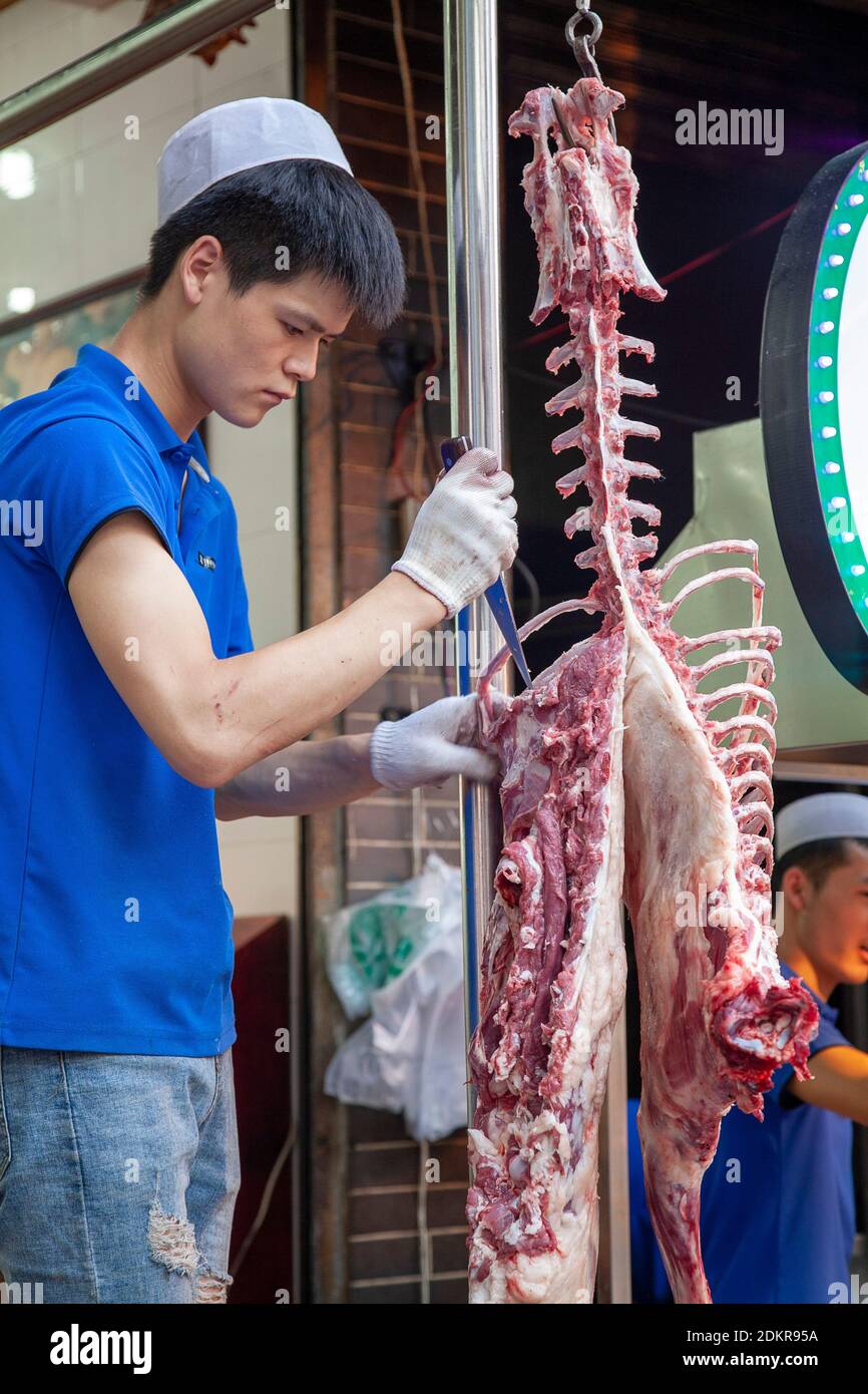 Chinese man carving meat from lamb carcass skeleton hanging for sale at street food vendor shop in Muslim Quarter Xian Xi'an ChinaMuslim Quarter Xian Stock Photo