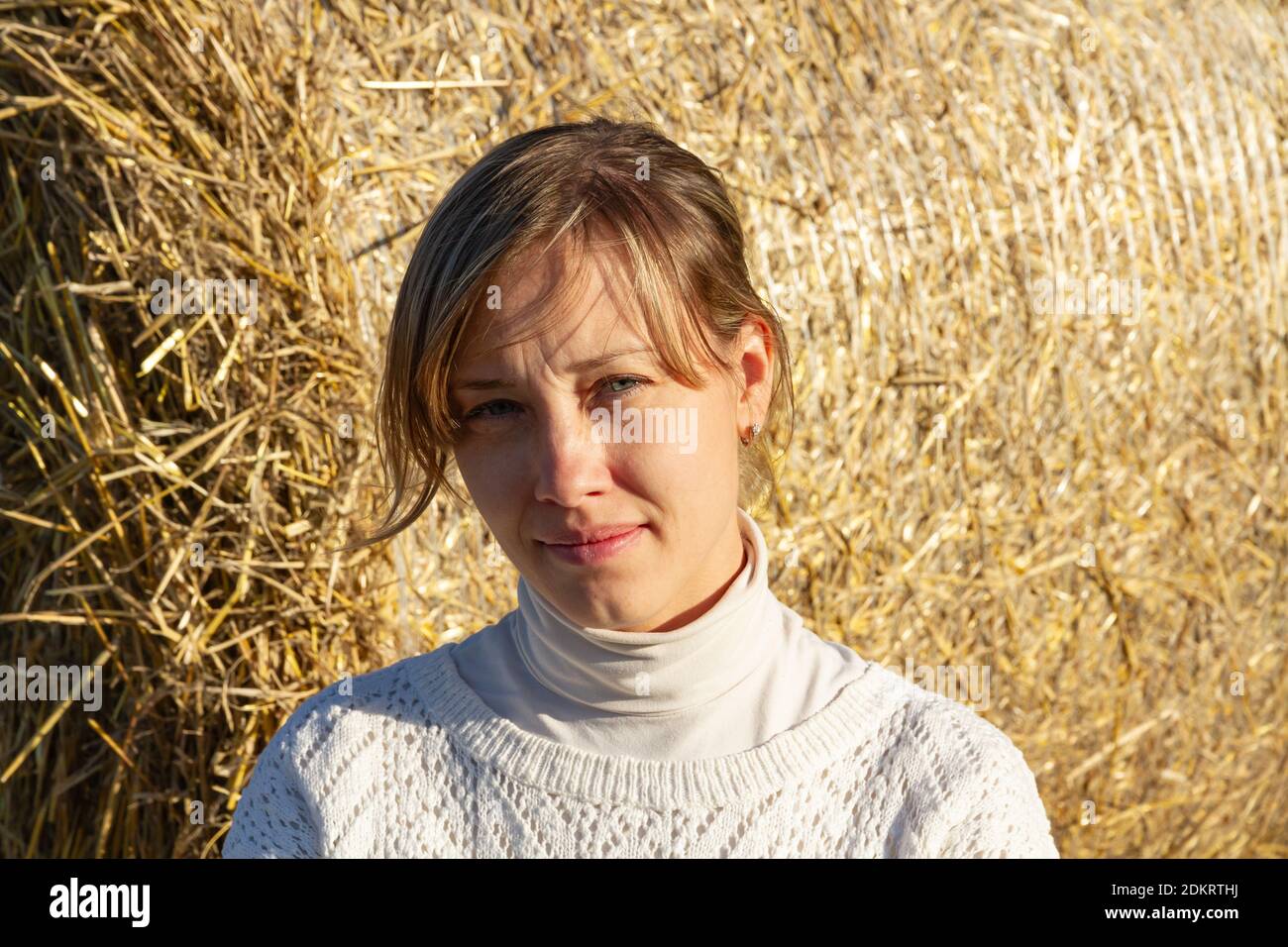 portrait of a young pretty girl in a white sweater on the background of a haystack, looking at the camera Stock Photo