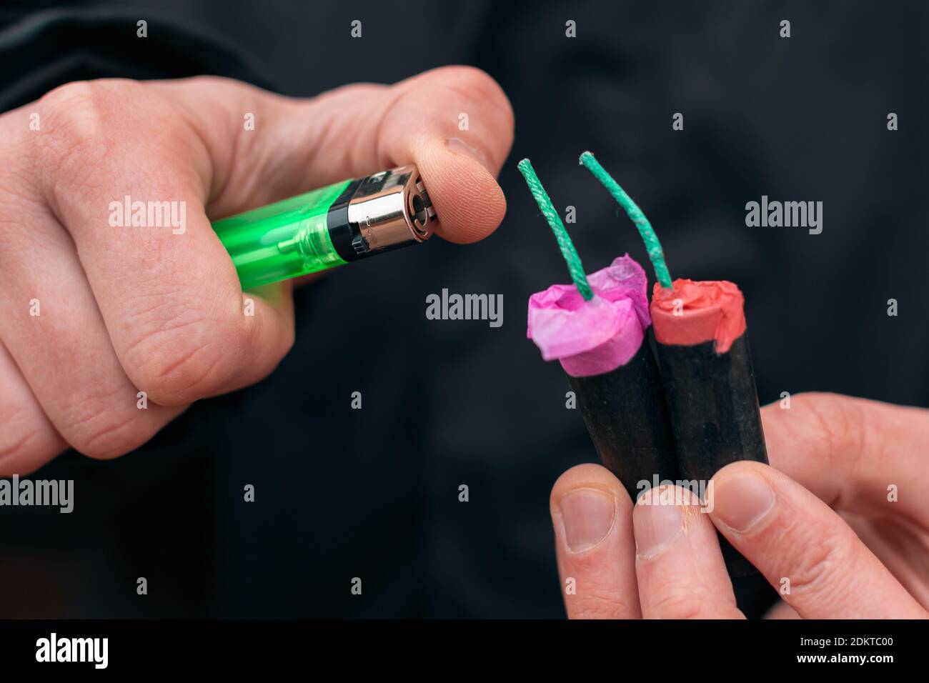 Setting Fire to the several Firecrackers. Man in Black Clothes Lighting Up Two Petards at the Same Time. Firing Up the Pyrotechnic with Green Gas Lighter Outdoors Stock Photo