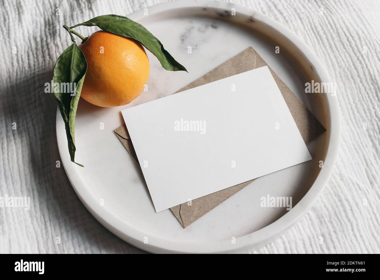 Summer wedding stationery mock-up scene. Blank greeting card, invitation. Craft envelope and orange fruit on elegant marble tray. White muslin Stock Photo