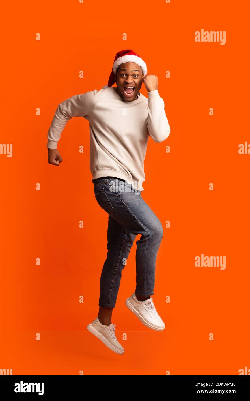 Ready For Party. Cheerful African Guy In Santa Hat Running In Air Stock Photo
