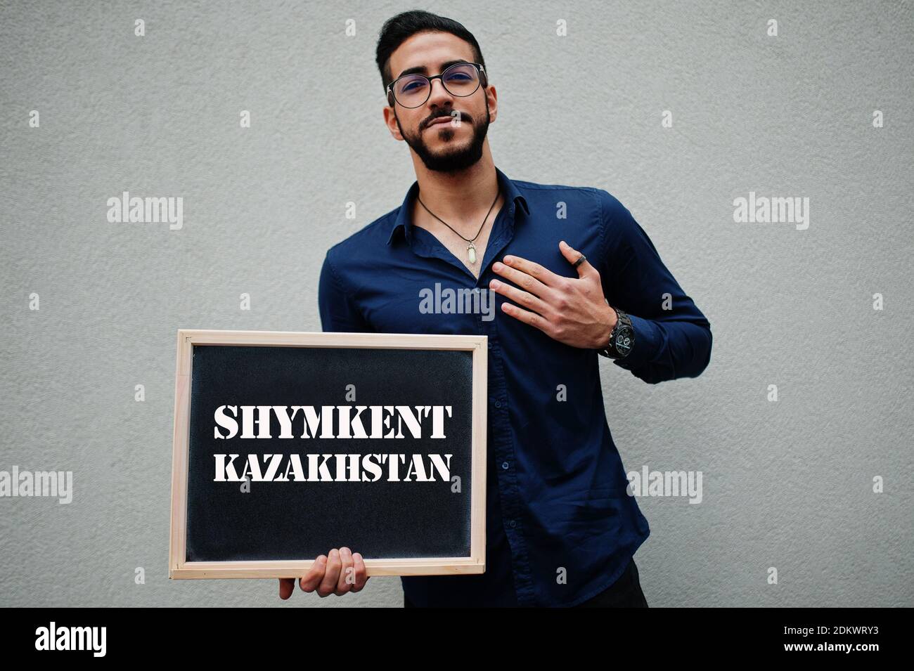 Arab man wear blue shirt and eyeglasses hold board with Shymkent Kazakhstan inscription. Largest cities in islamic world concept. Stock Photo