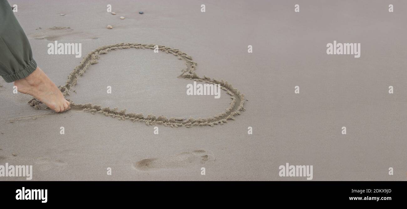 Unrecognizable woman draws a heart on the sand with her foot on 14th February. Valentine's Day 2021. Stock Photo
