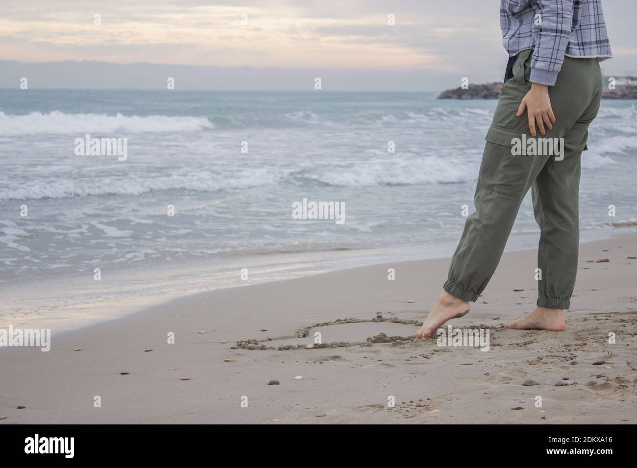 Horizontal conceptual image with an ocean background. Unrecognizable woman draws a heart on the sand with her foot on 14th February. Valentine's Day. Stock Photo