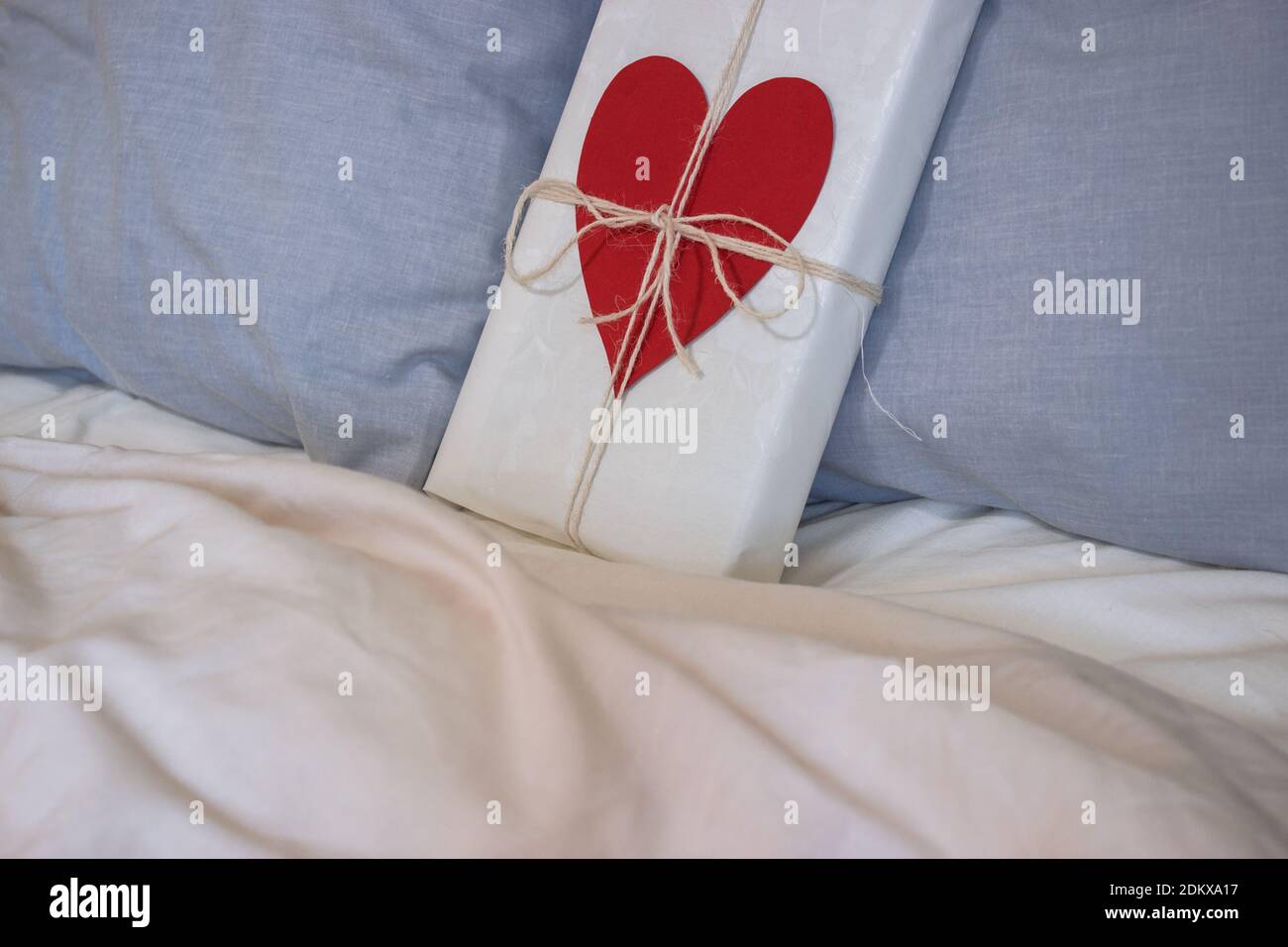 A gift with a ribbon and a heart is placed on the bed of a loving couple's house for Valentine's Day. Valentine's present. 14th February 2021. Stock Photo