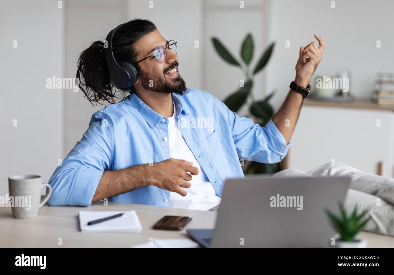 Break at work. Joyful african american businessman listening music in headphones and playing virtual guitar, relaxing at workplace in office. Stock Photo