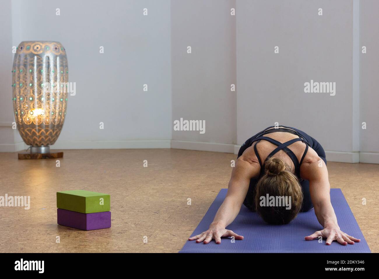Yogi woman practices child pose in studio with lamp by white background. Female yoga student in balasana on purple mat with props on side Stock Photo
