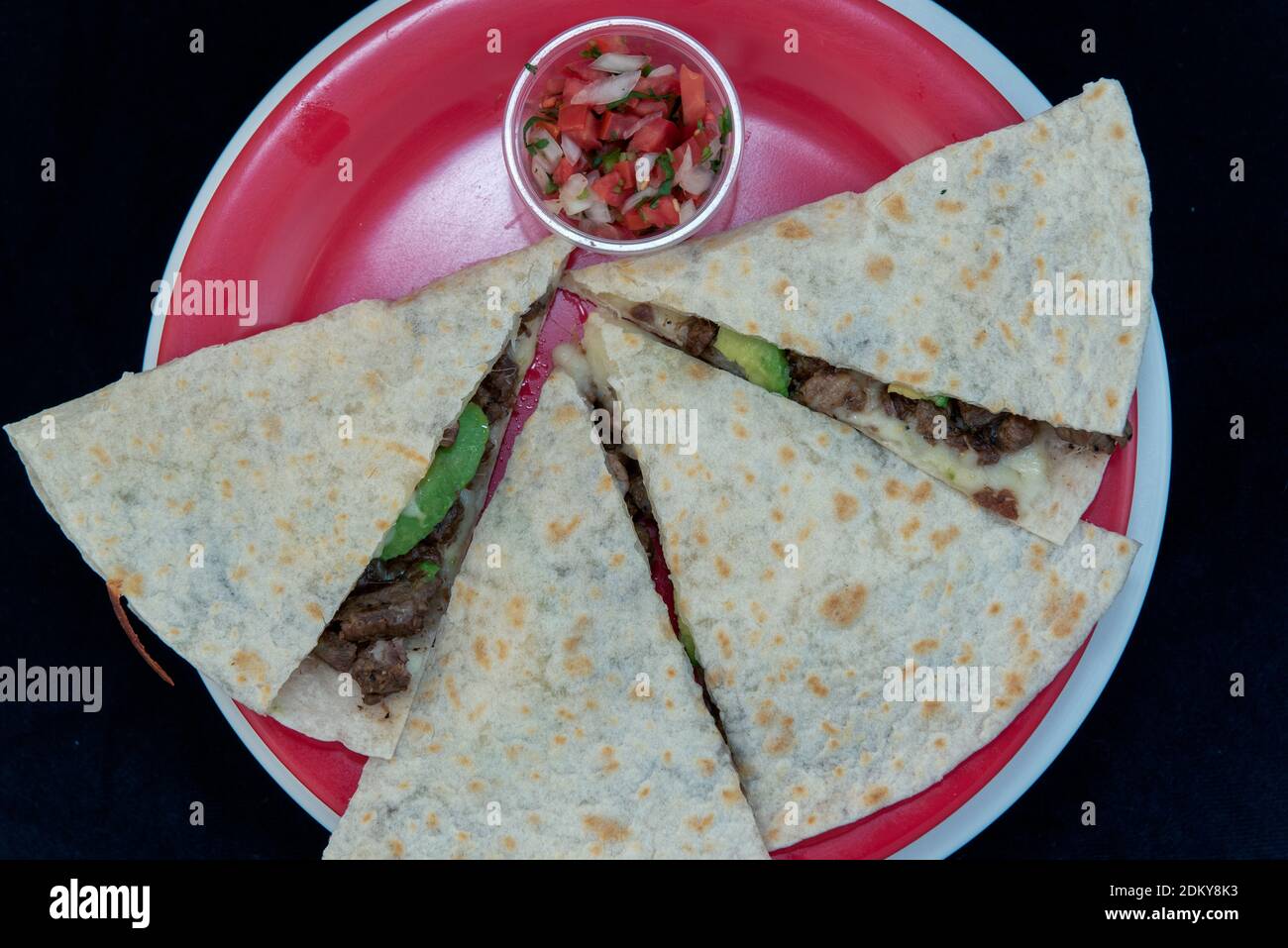 Overhead view of steak Asada quesadilla loaded with fresh meat, melted cheese and cut in wedges and presented on a red plate. Stock Photo