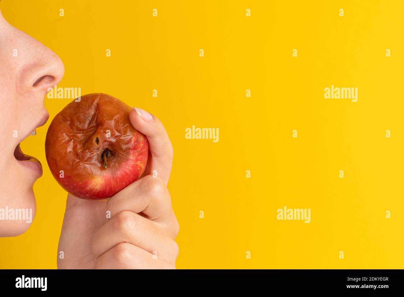 The girl bites a rotten apple with a worm on a yellow background. Expired products, junk food. Stock Photo