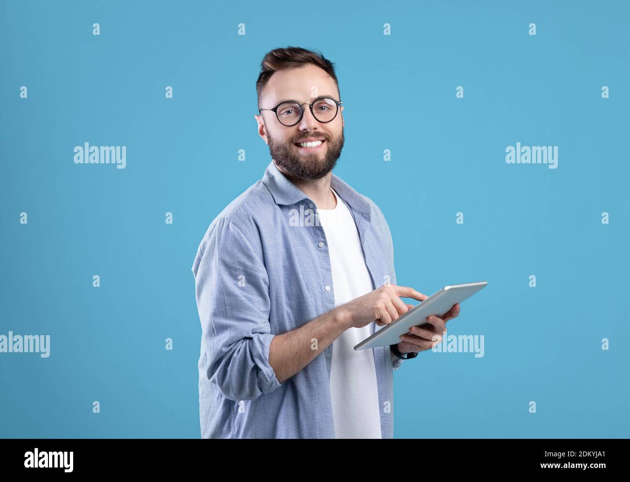 Positive young guy in casual wear using tablet computer over blue studio background Stock Photo