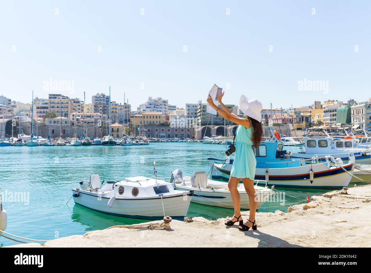 Smiling stylish girl at the Venetian port of Heraklion Crete Greece. Elegant girl in turquoise dress, sunhat map and camera on walking sightseeing tri Stock Photo
