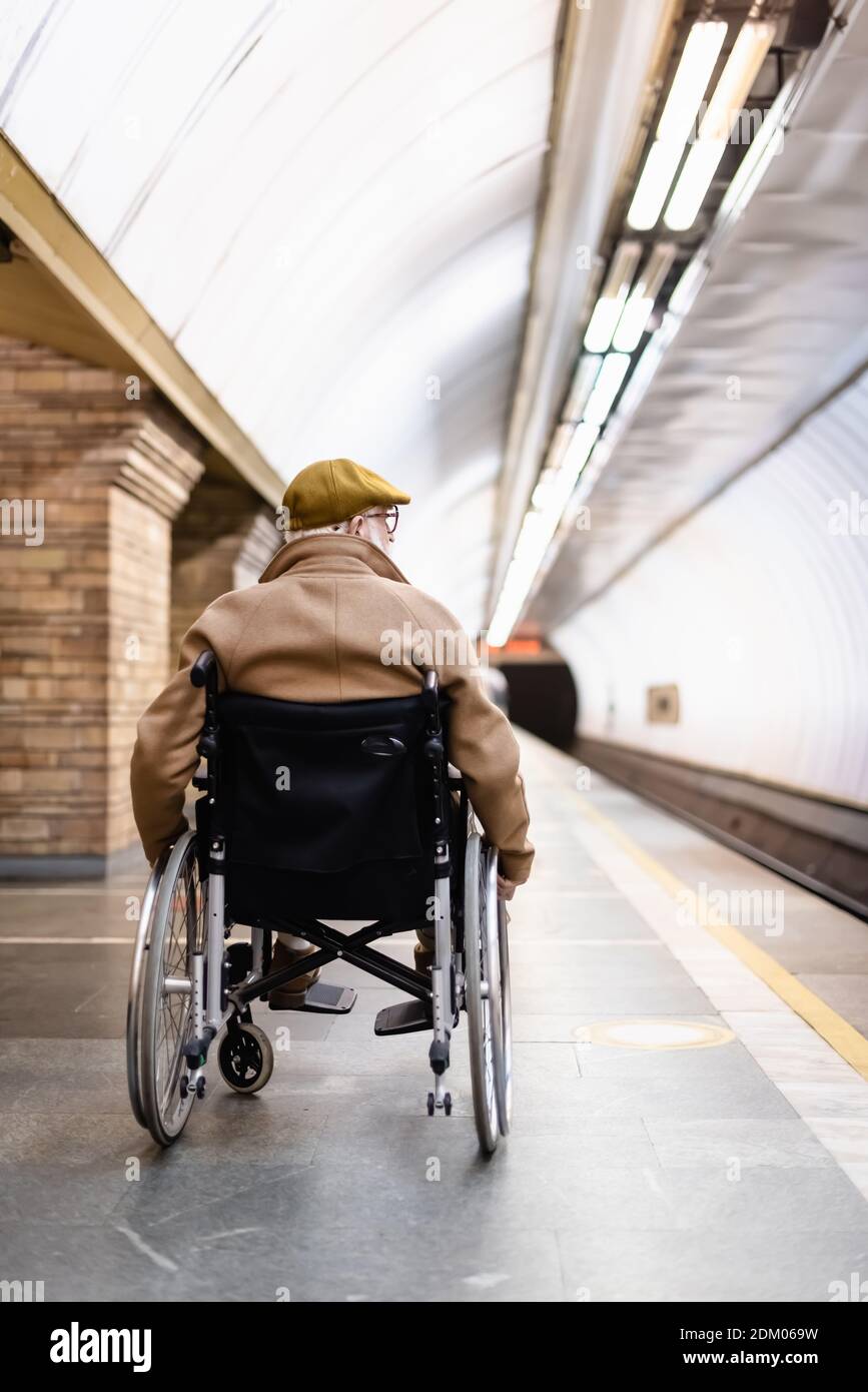 back view of senior man in wheelchair, wearing coat and cap, on platform of metro station Stock Photo